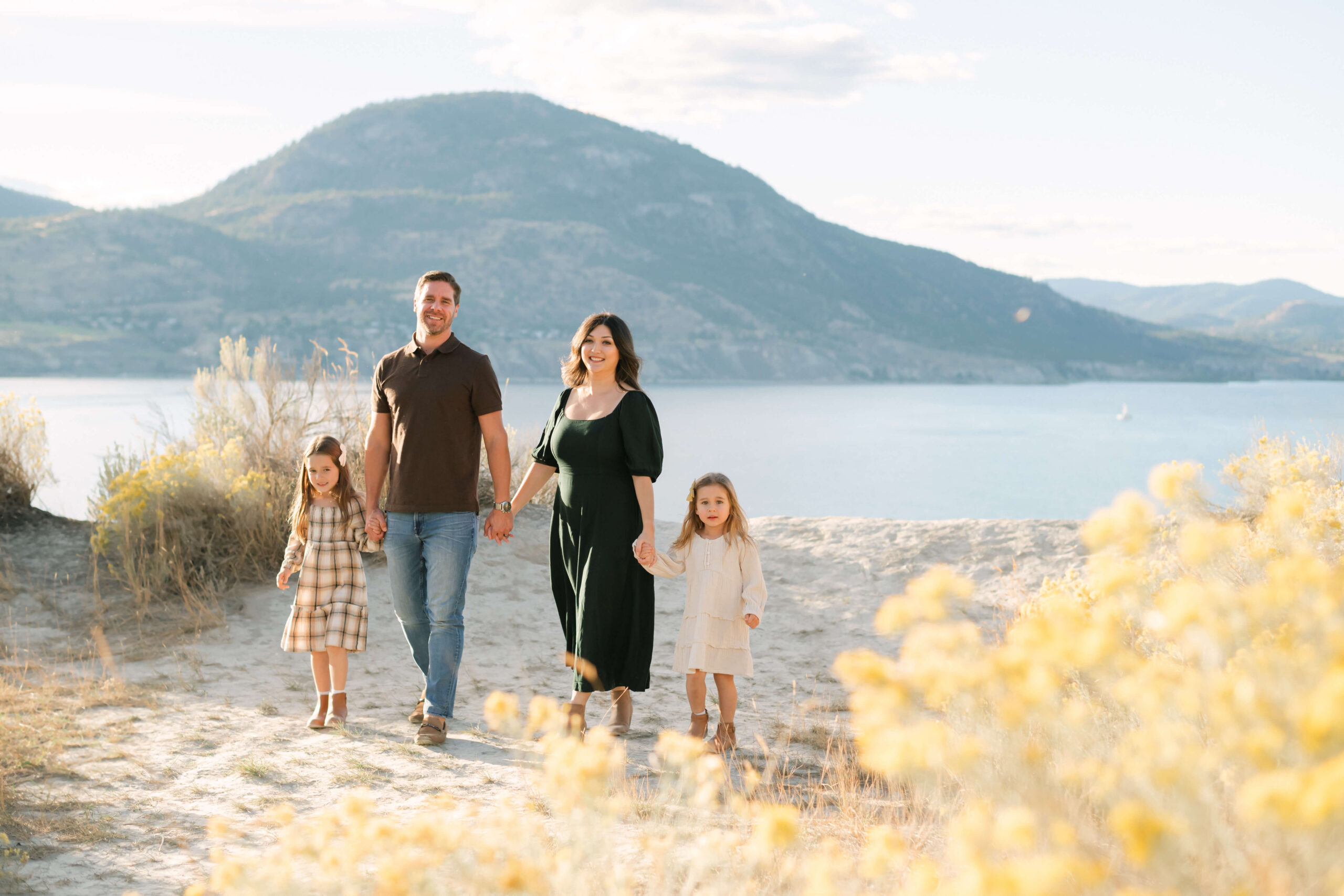 A beautiful family of 4 (dad, mom, two girls around 4 and 6) walk in a field of sagebrush overlooking Okanagan Lake and the mountains of Penticton
