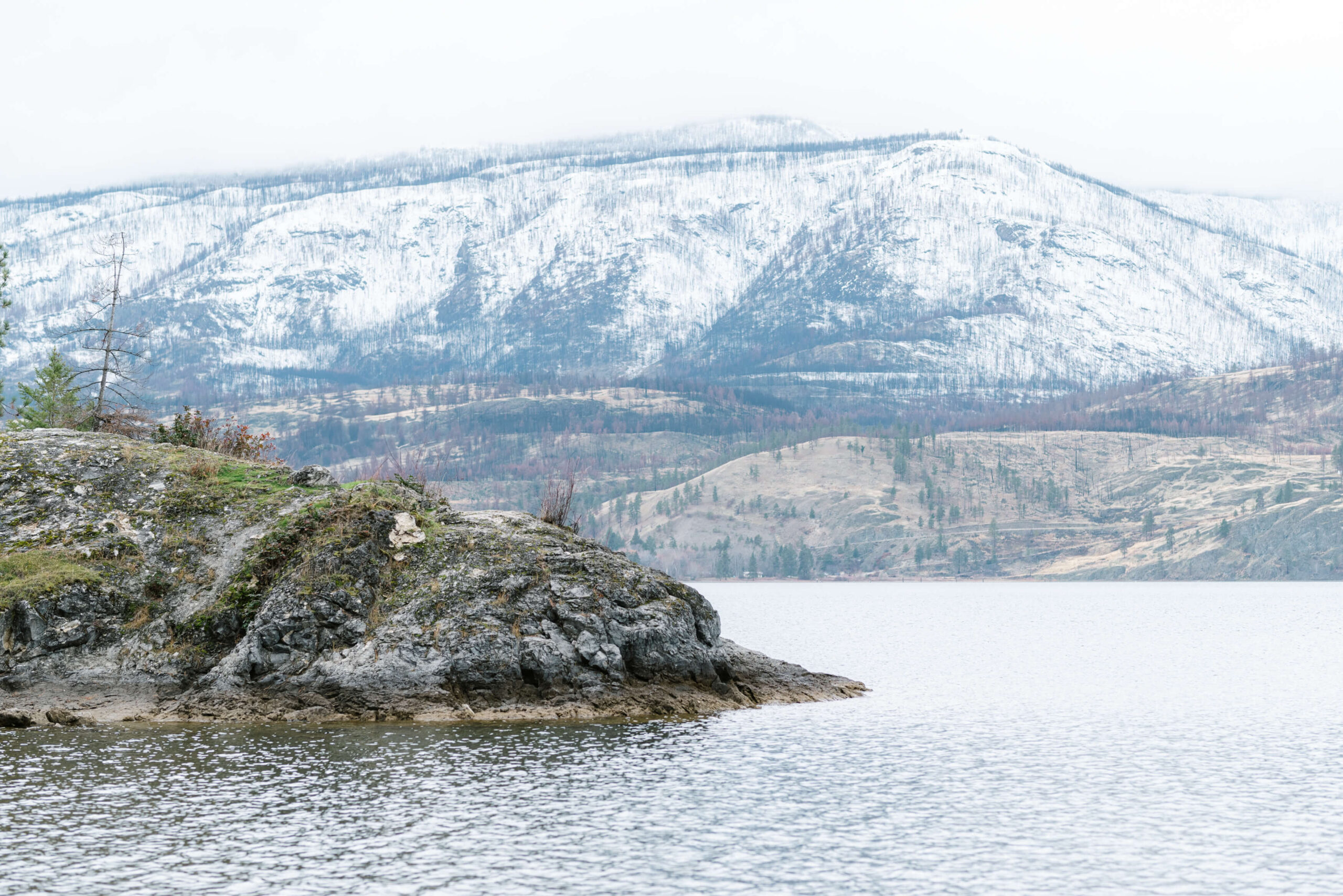 Okanagan ski mountain covered in snow in the background, the lake is in the foreground with a rock area visible on the left hand side.