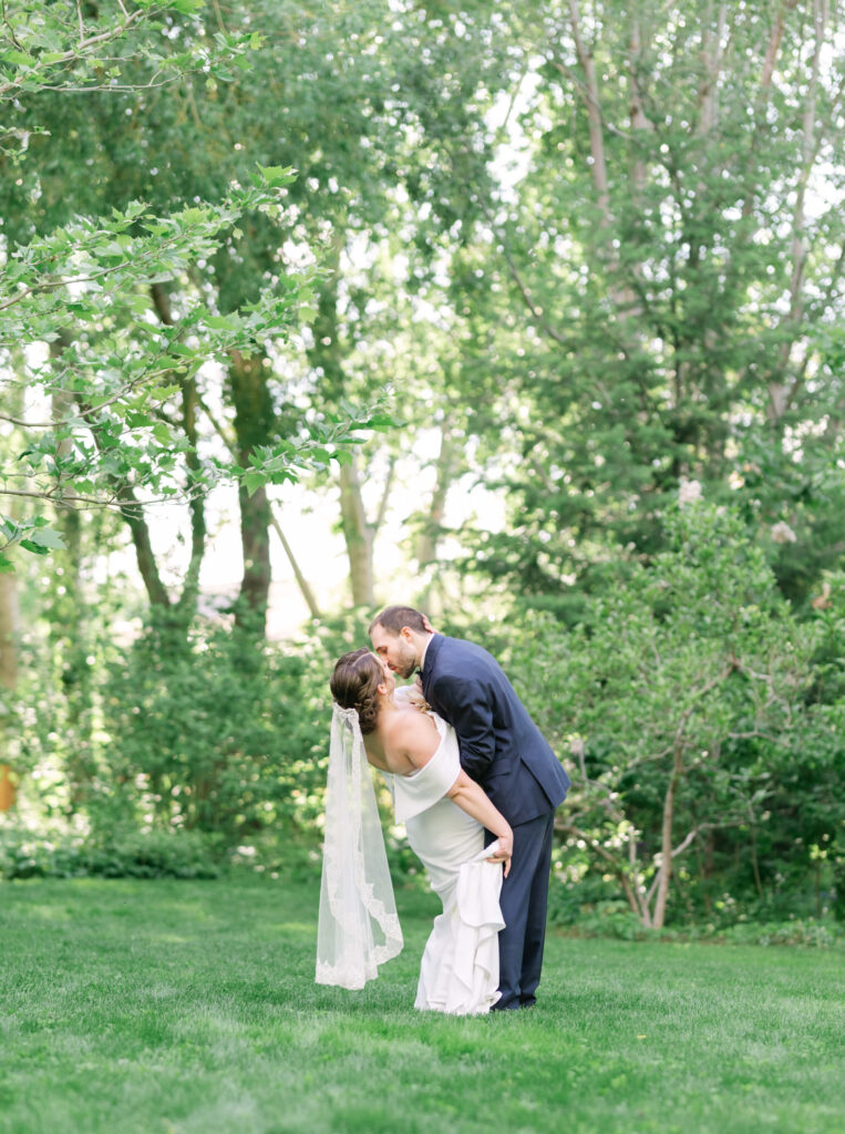 bride and groom kiss in penticton linden gardens wedding venue, background is a grass lawn with trees and sun shining through