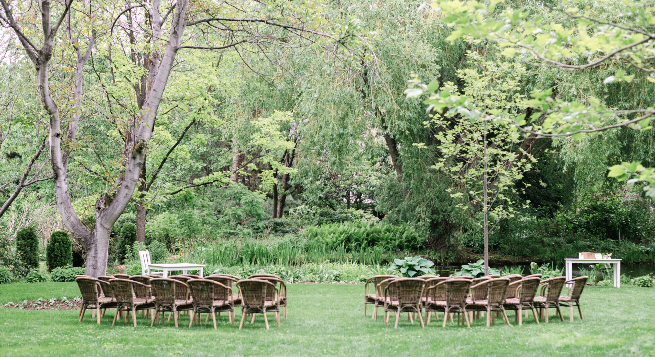 wicker chairs set up for a wedding ceremony on green grass with treed garden in the background